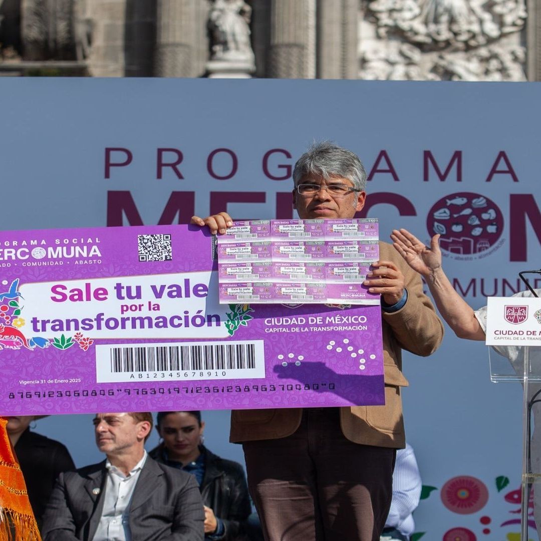 Una persona adulta mayos sostiene un vale en un evento junto a la jefa de gobierno, Clara Brugada.