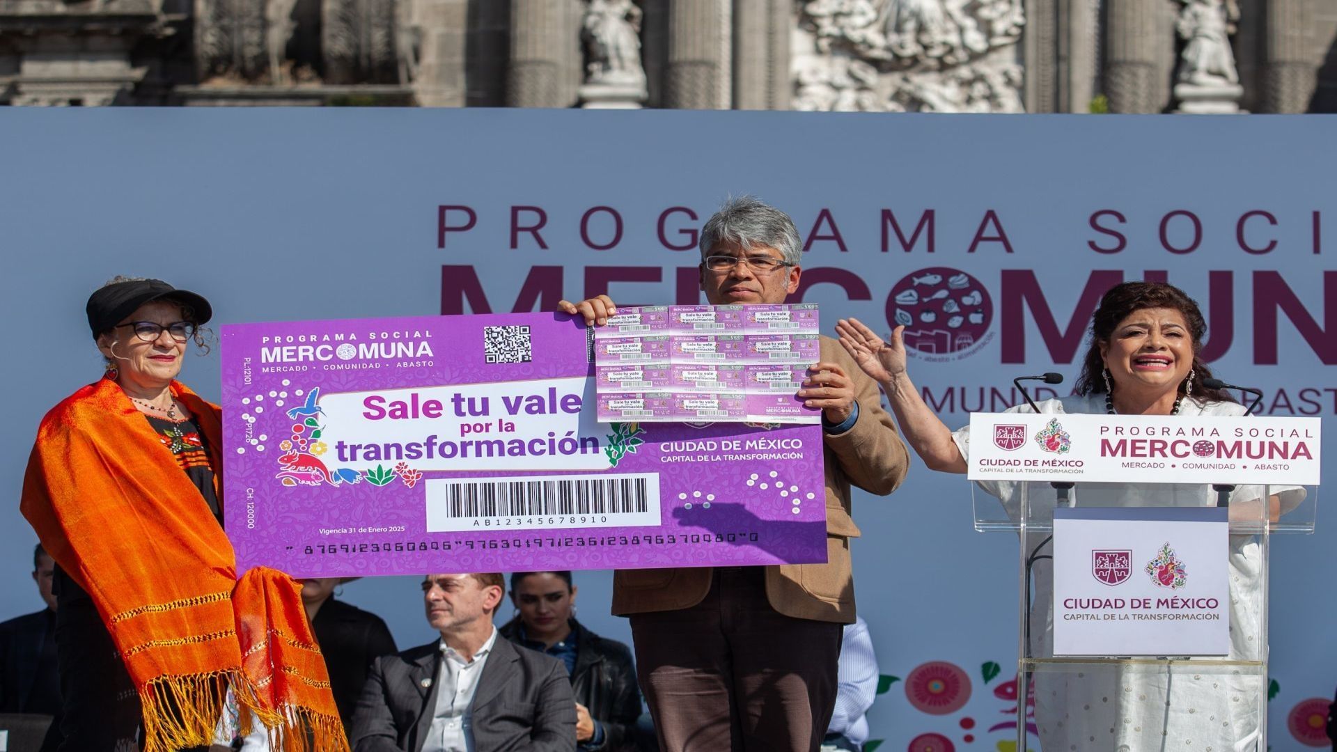 Una persona adulta mayos sostiene un vale en un evento junto a la jefa de gobierno, Clara Brugada.