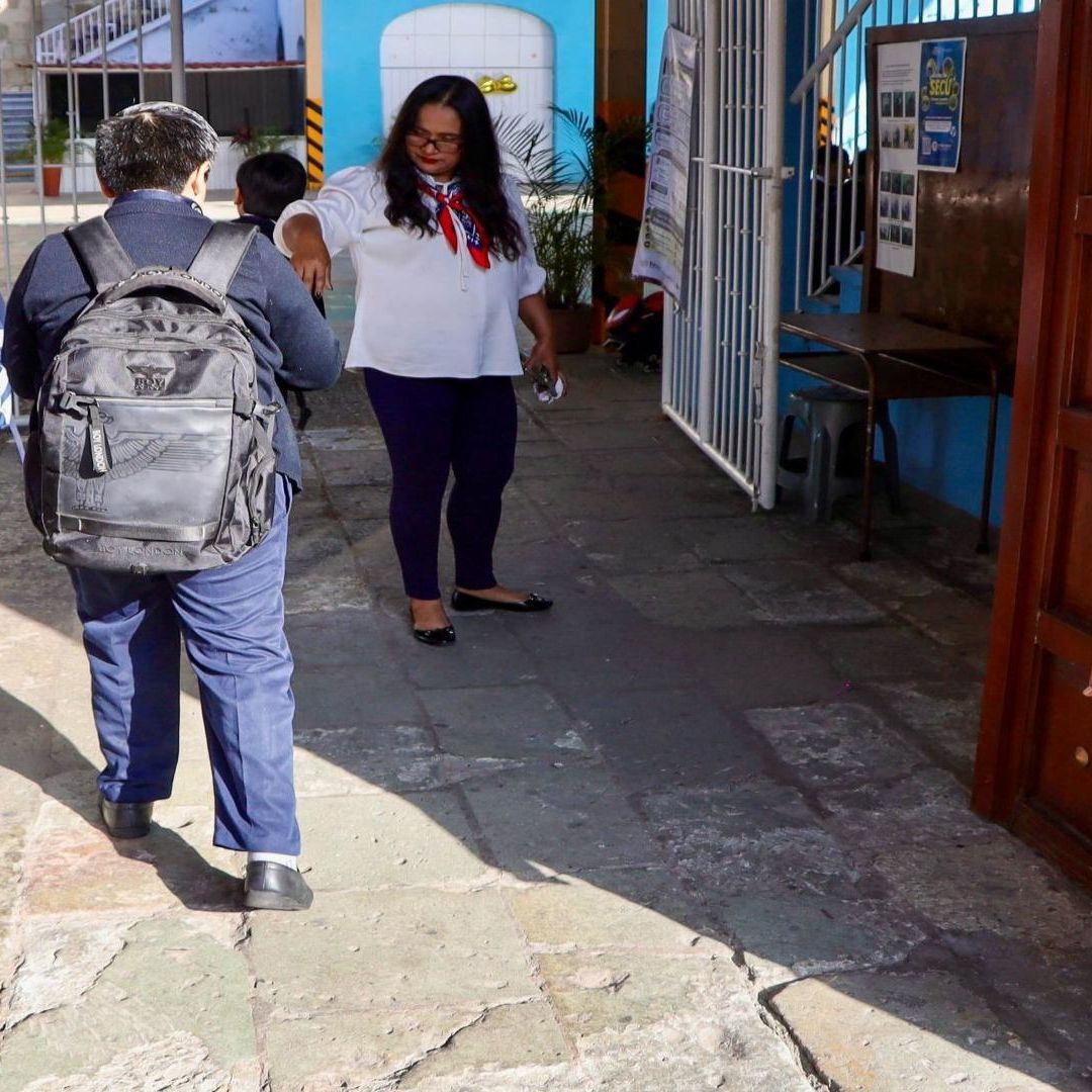Estudiantes llevan una mochila, mientras los reciben en la entrada de la escuela.