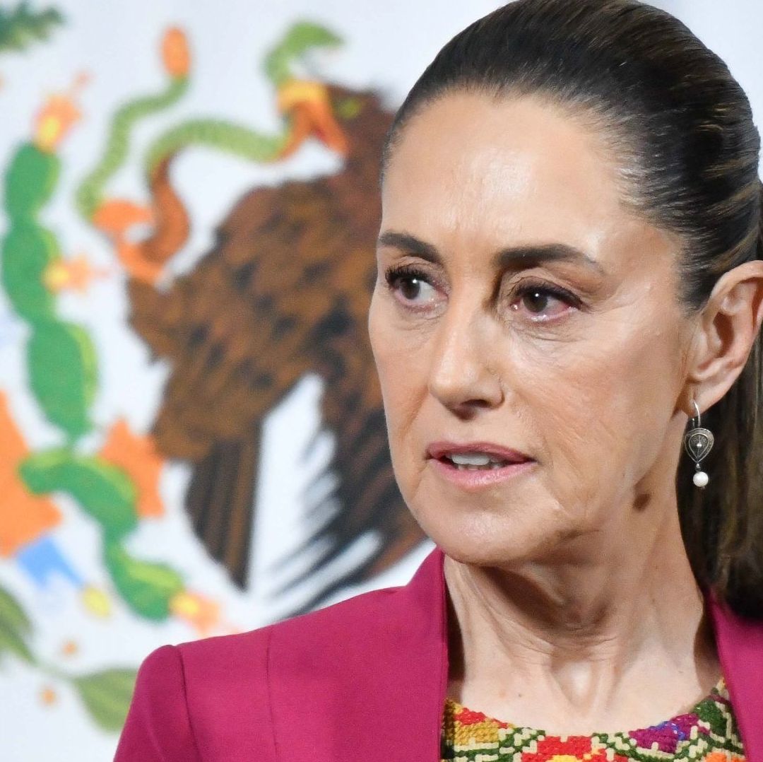 La presidenta Claudia Sheinbaum viste un raje de color rosa y se encuentra al lado de una bandera.