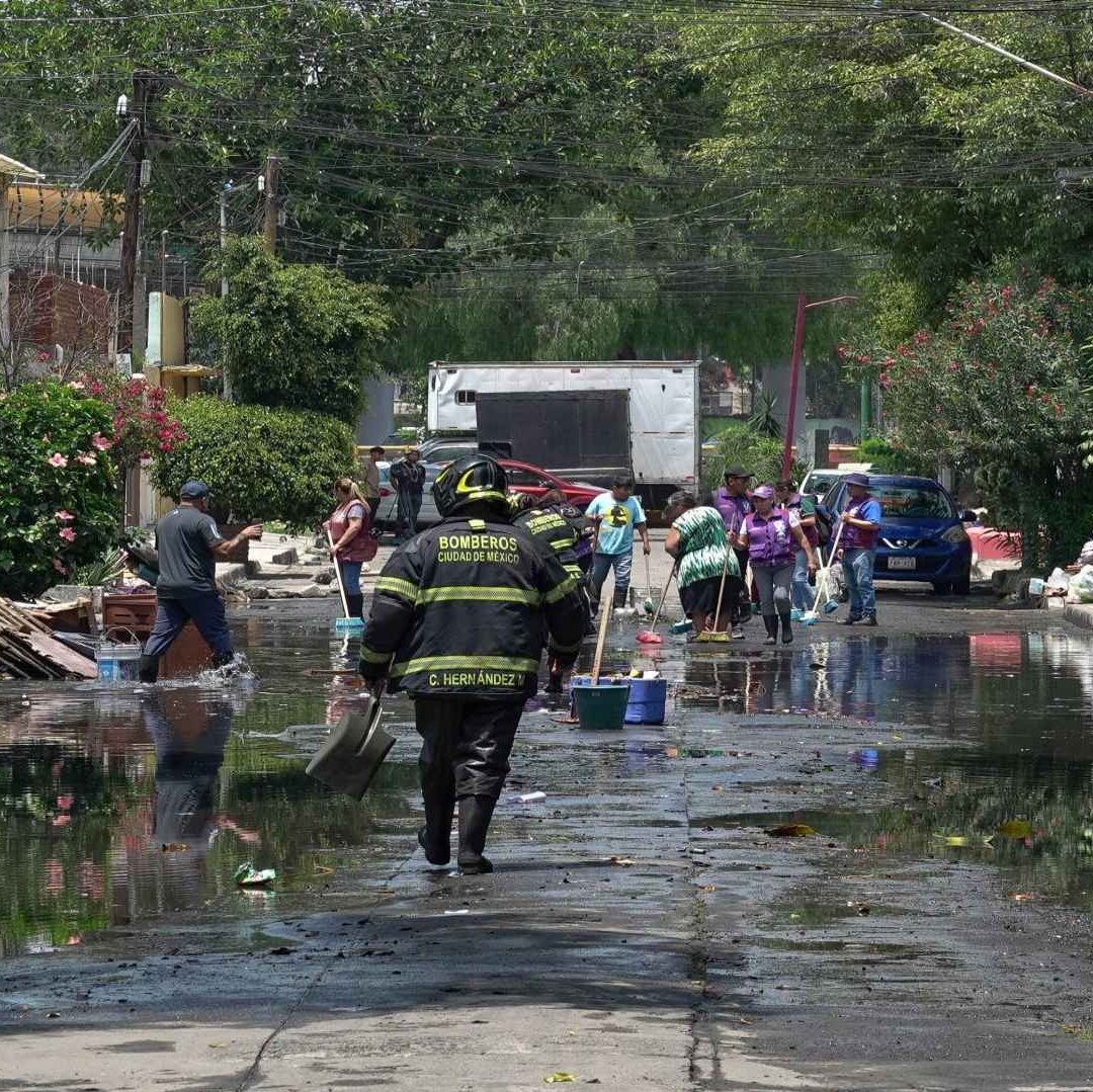 Un bombero caminado en medio de una calle encharcada, a su alrededor, personas afectadas por las lluvias realizan labores de limpieza.