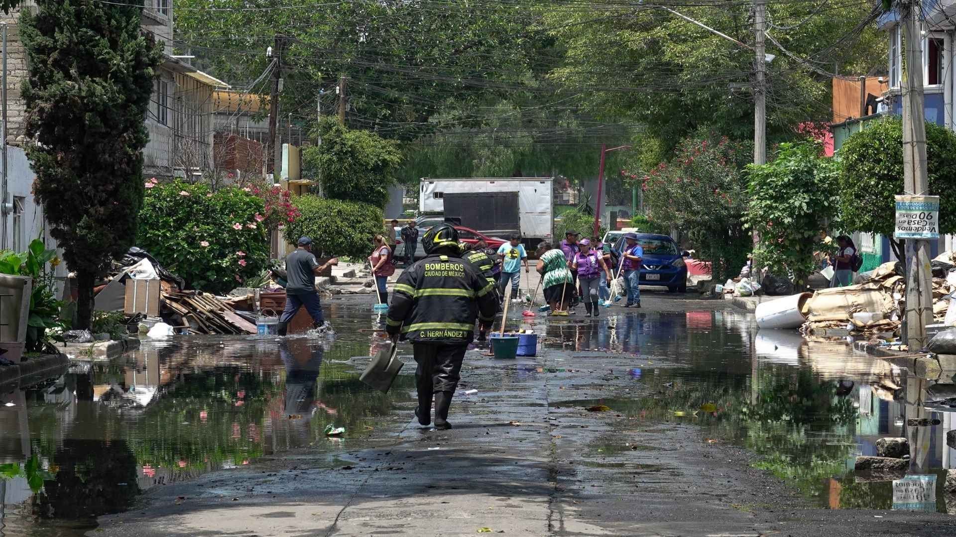 Un bombero caminado en medio de una calle encharcada, a su alrededor, personas afectadas por las lluvias realizan labores de limpieza.