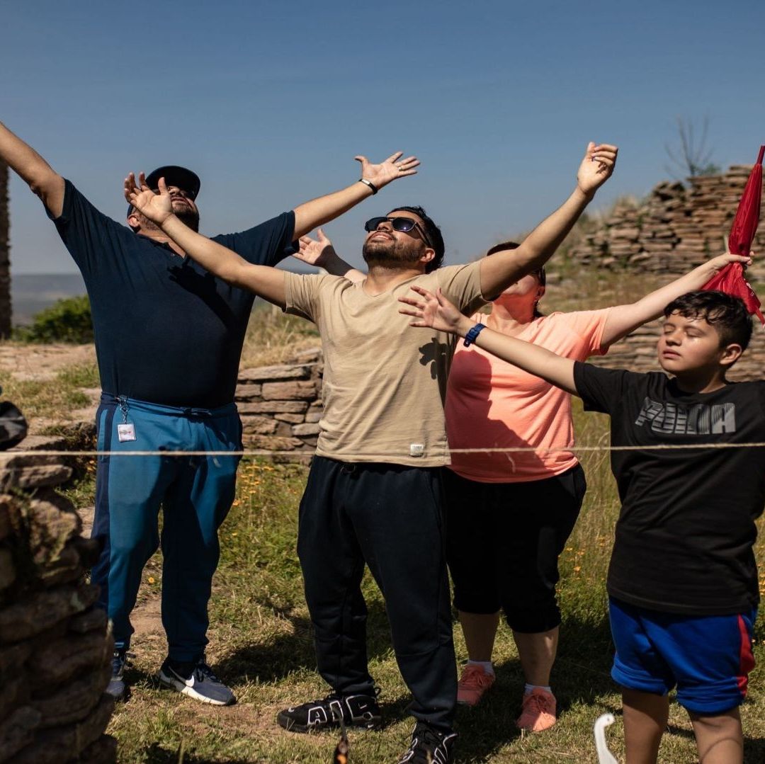 Cuatro personas se encuentran en un espacio verde, expandiendo lo brazos y volteando hacia el cielo.
