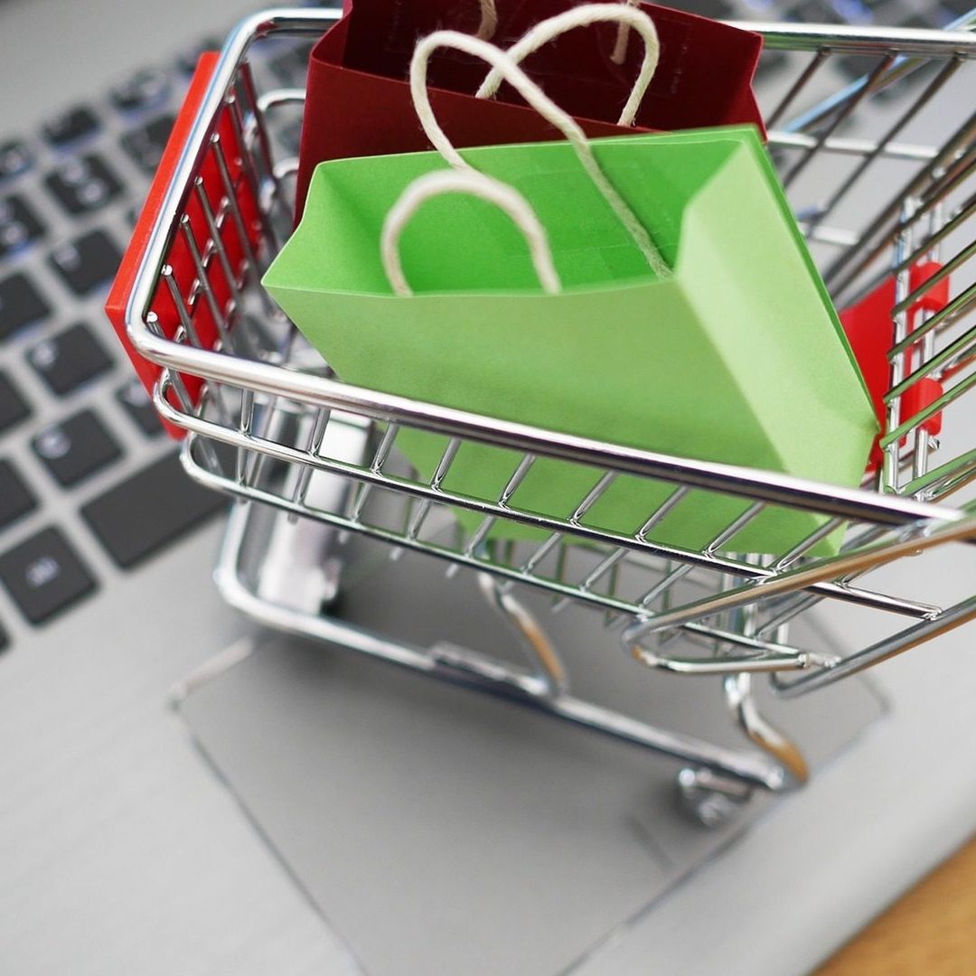 Un minicarrito de supermercado con bolsas pequeñas de colores, arriba de una laptop.