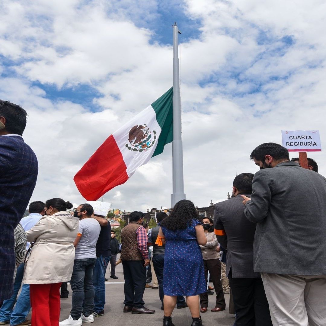 Un hombre de traje junto a más personas, se encuentran cerca de una bandera de México.