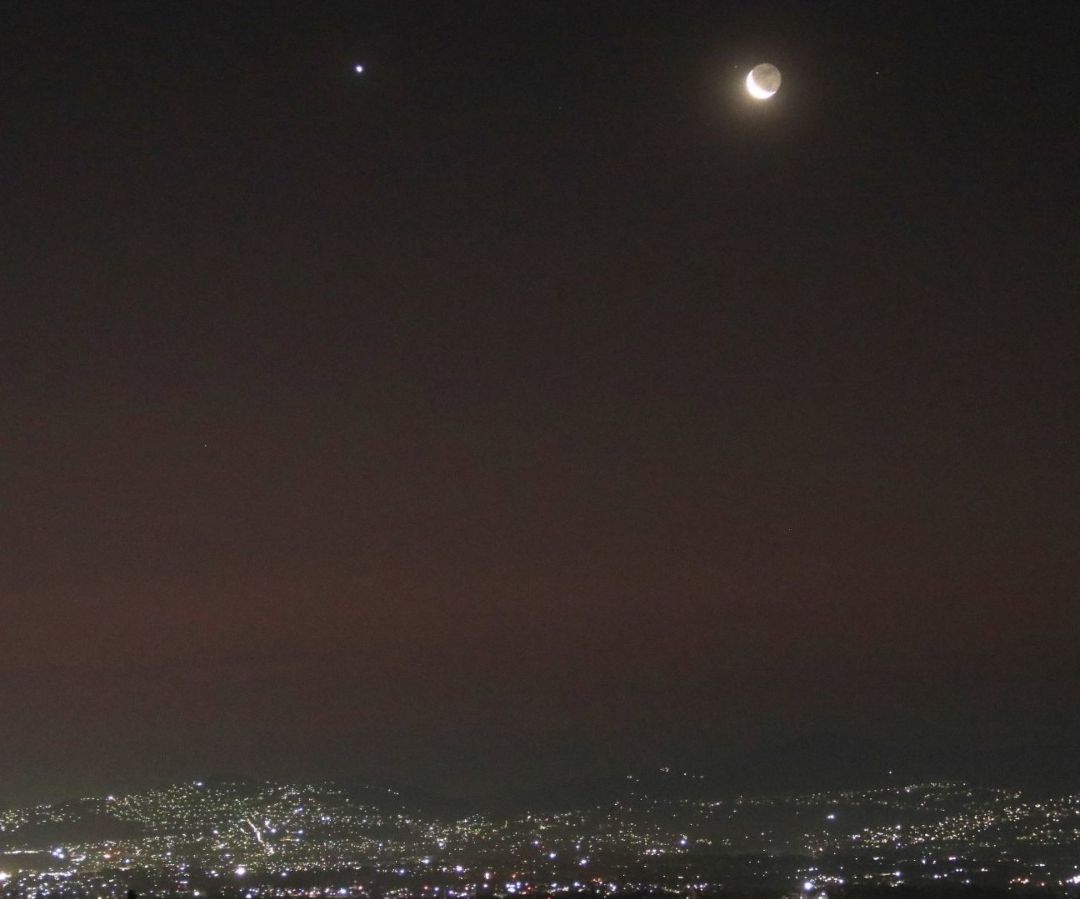 El cielo nocturno con varias luces de las casas, se puede ver la Luna y un planeta.