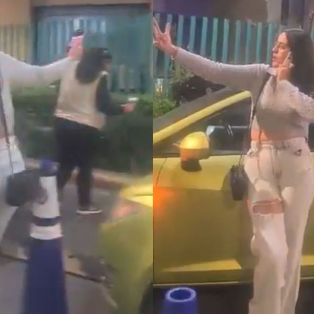 Una mujer está recargada en auto de color verde, mientras usa su celular para grabar.