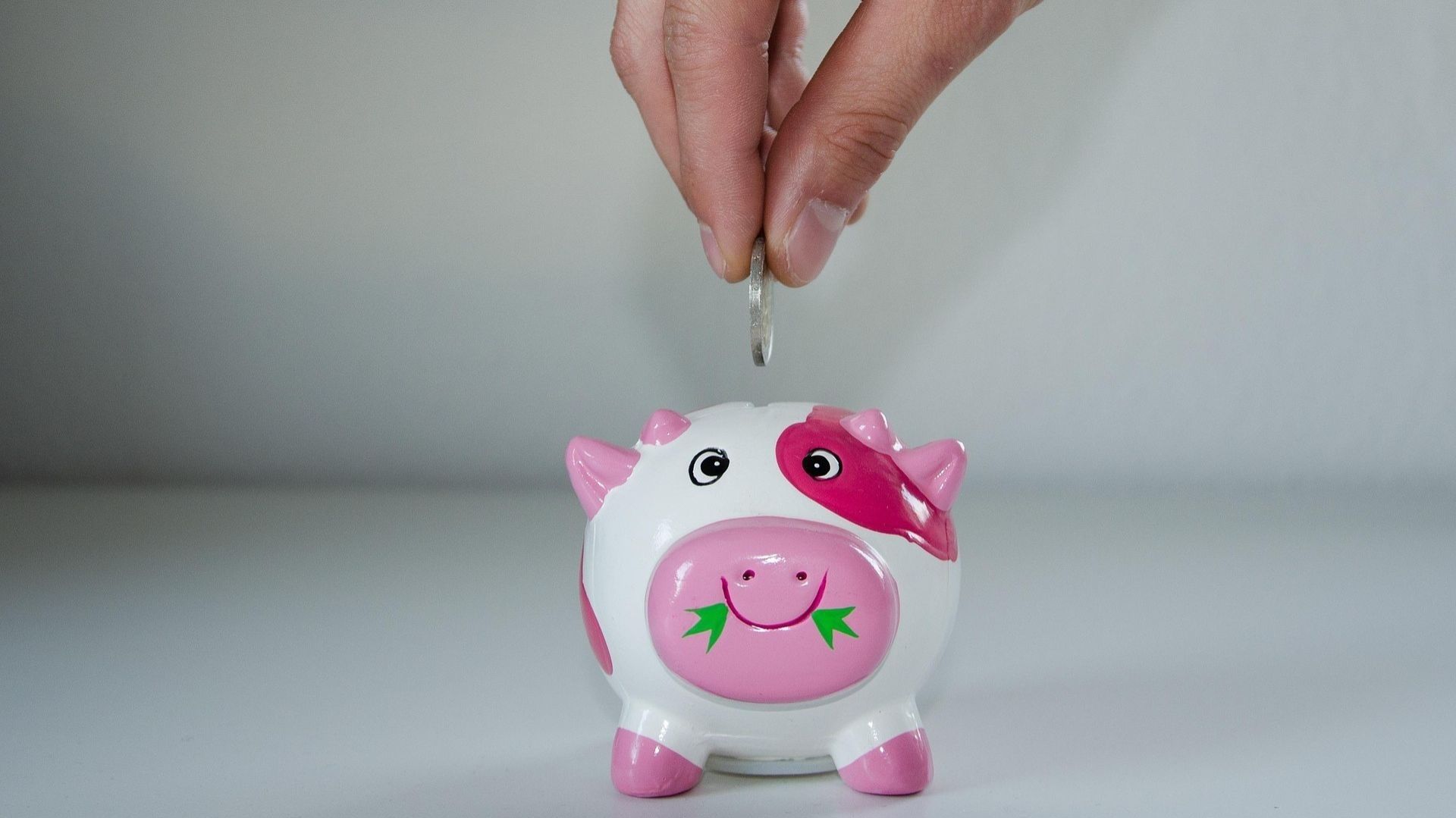 Una alcancía de puerquito de color rosa, una mano deposita una moneda.