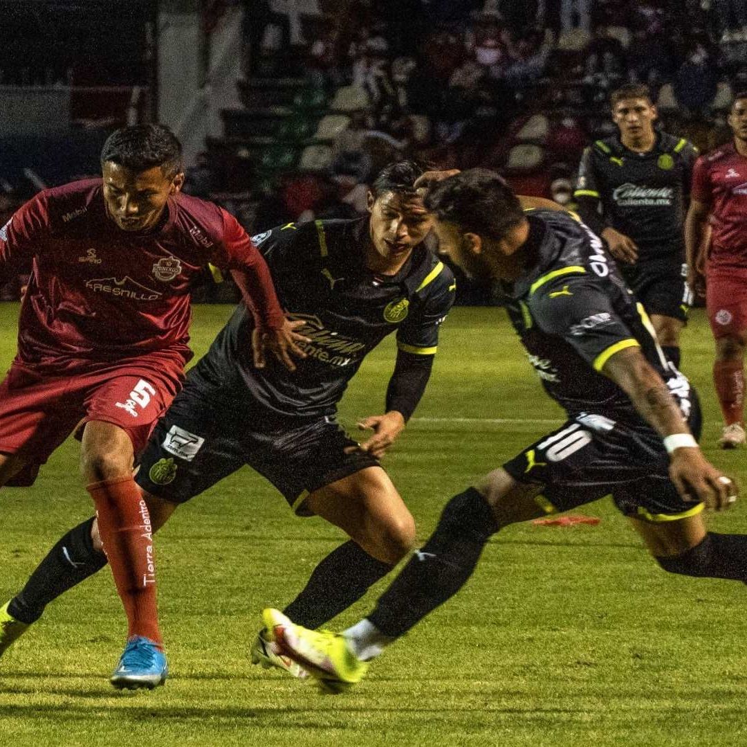 Jugadores de futbol portan uniformes negros y vino; se disputan el balón.