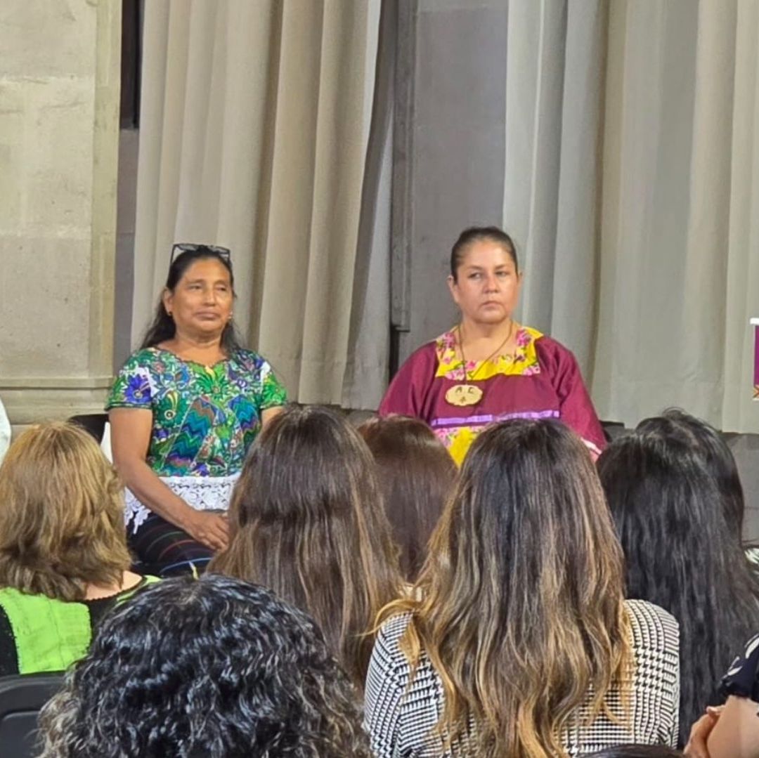 Un hombre vestido de blanco habla en un micrófono. Hay mujeres vestidas con trajes típicos de México.