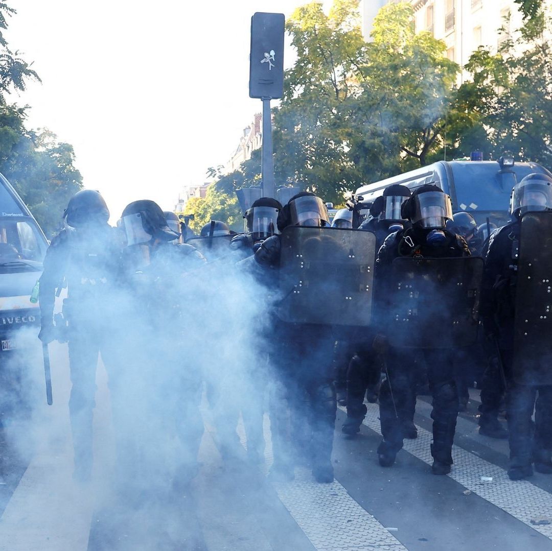 Humo en medio de hombres vestidos de negro, quienes se protegen con un escudo de la Policía.