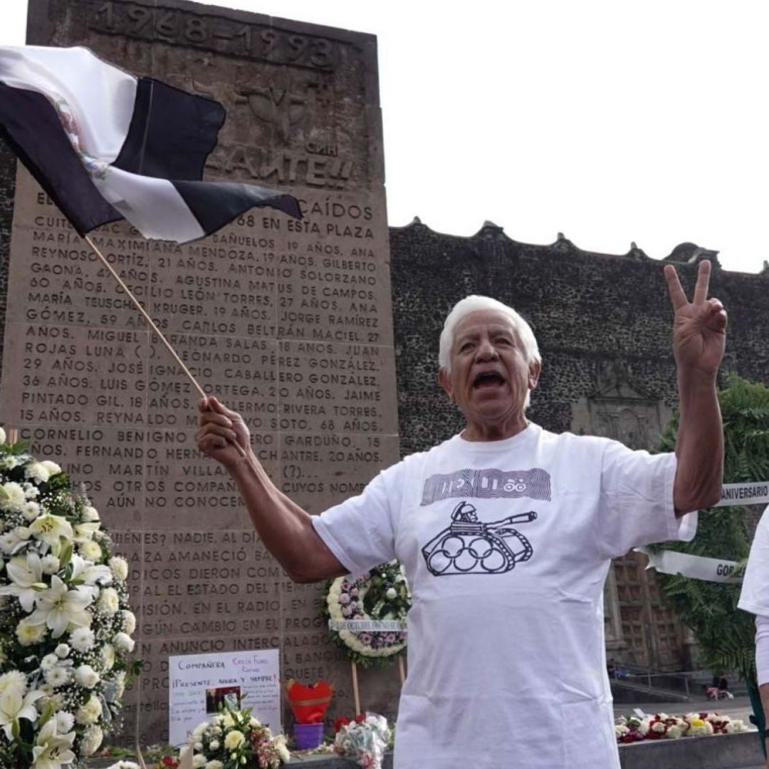 Dos personas adultas mayores tienen una bandera de color negro, detrás hay un memorial con letras.