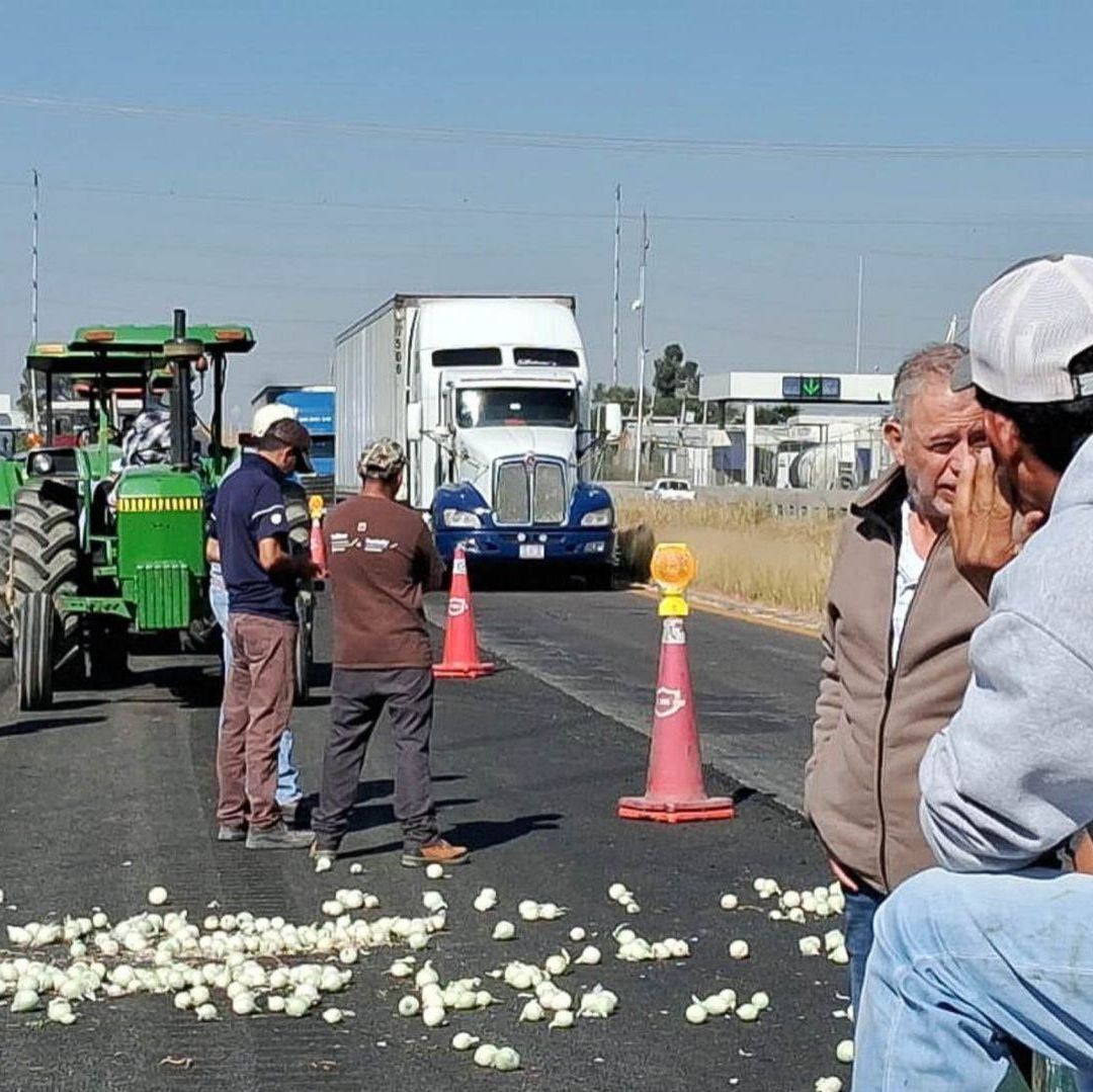 Varias personas sentadas, bloquean con camiones la carretera.