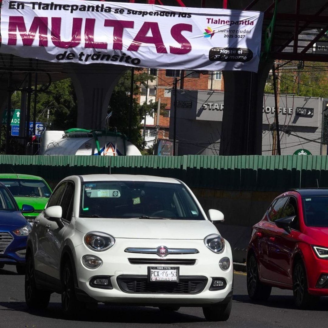 Carros en una vialidad, detrás un cártel que dice multas.