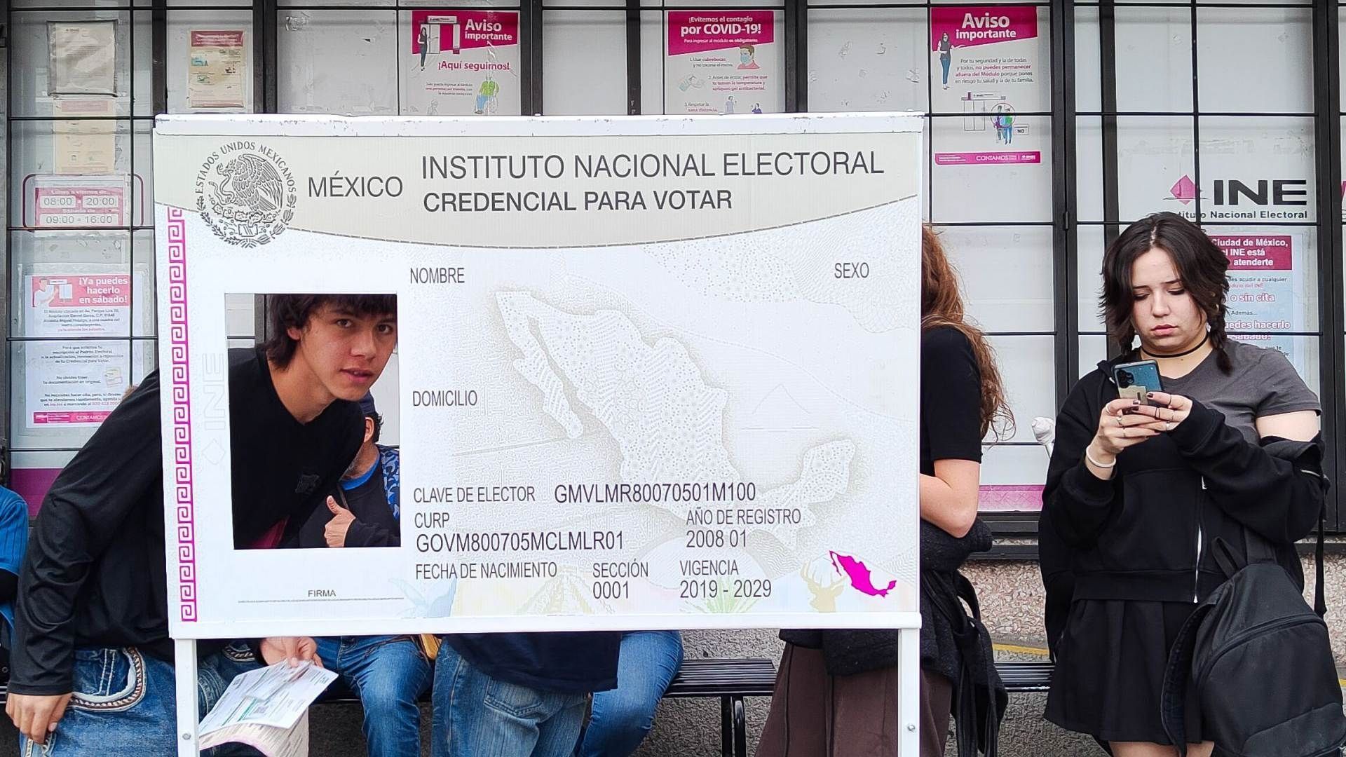 Ciudadanos esperan su turno en módulo del INE en Ciudad de México con una credencial gigante