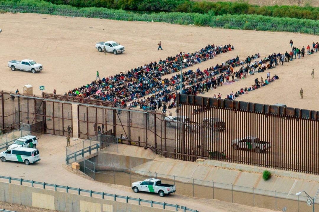 Grupo de migrantes esperando en la frontera entre México y Estados Unidos, con vehículos y vallas de seguridad presentes.