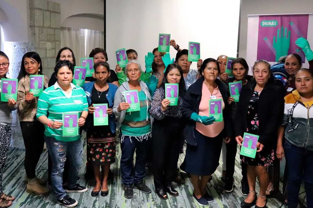 Grupo de mujeres sosteniendo folletos verdes con un símbolo de mano, junto a un banner "DIGNAS