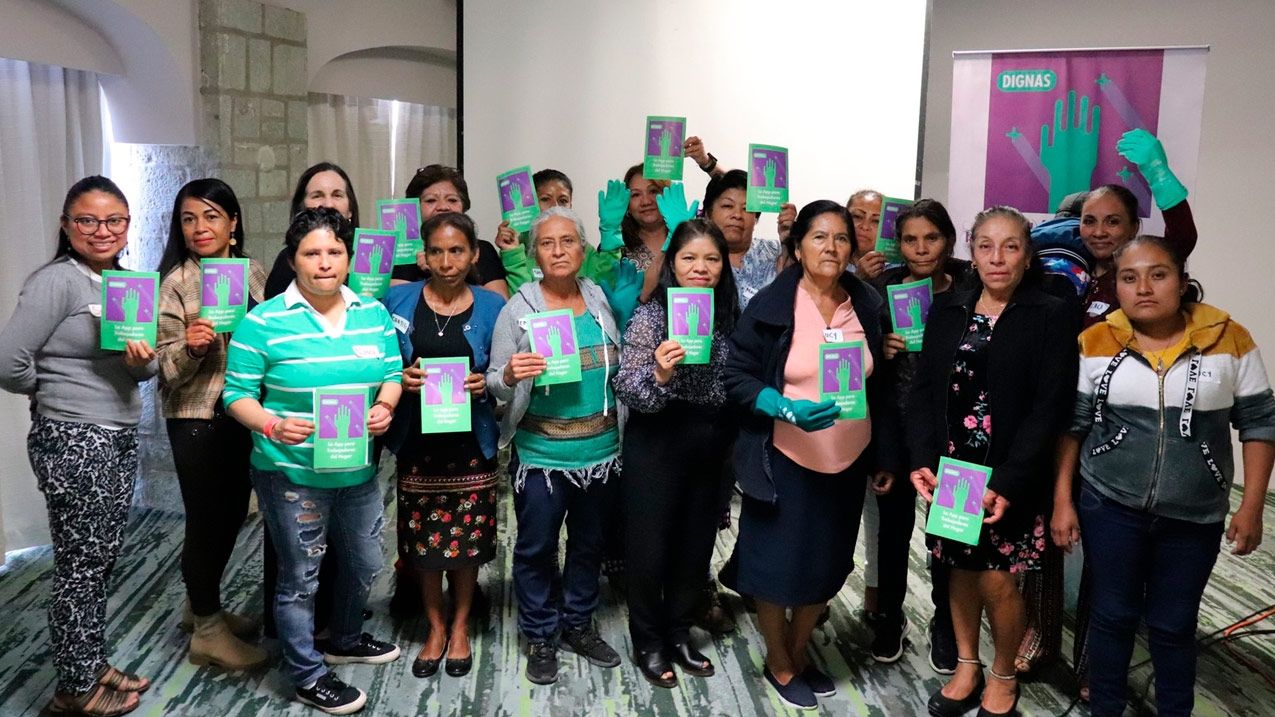 Grupo de mujeres sosteniendo folletos verdes con un símbolo de mano, junto a un banner "DIGNAS