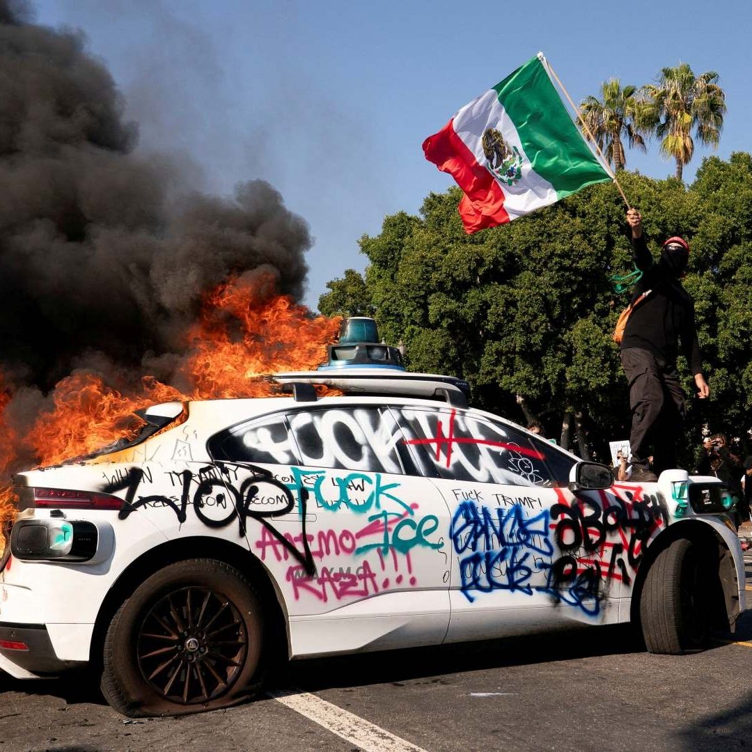 Manifestante ondea Bandera de México sobre un auto incendiado, en Los Ángeles