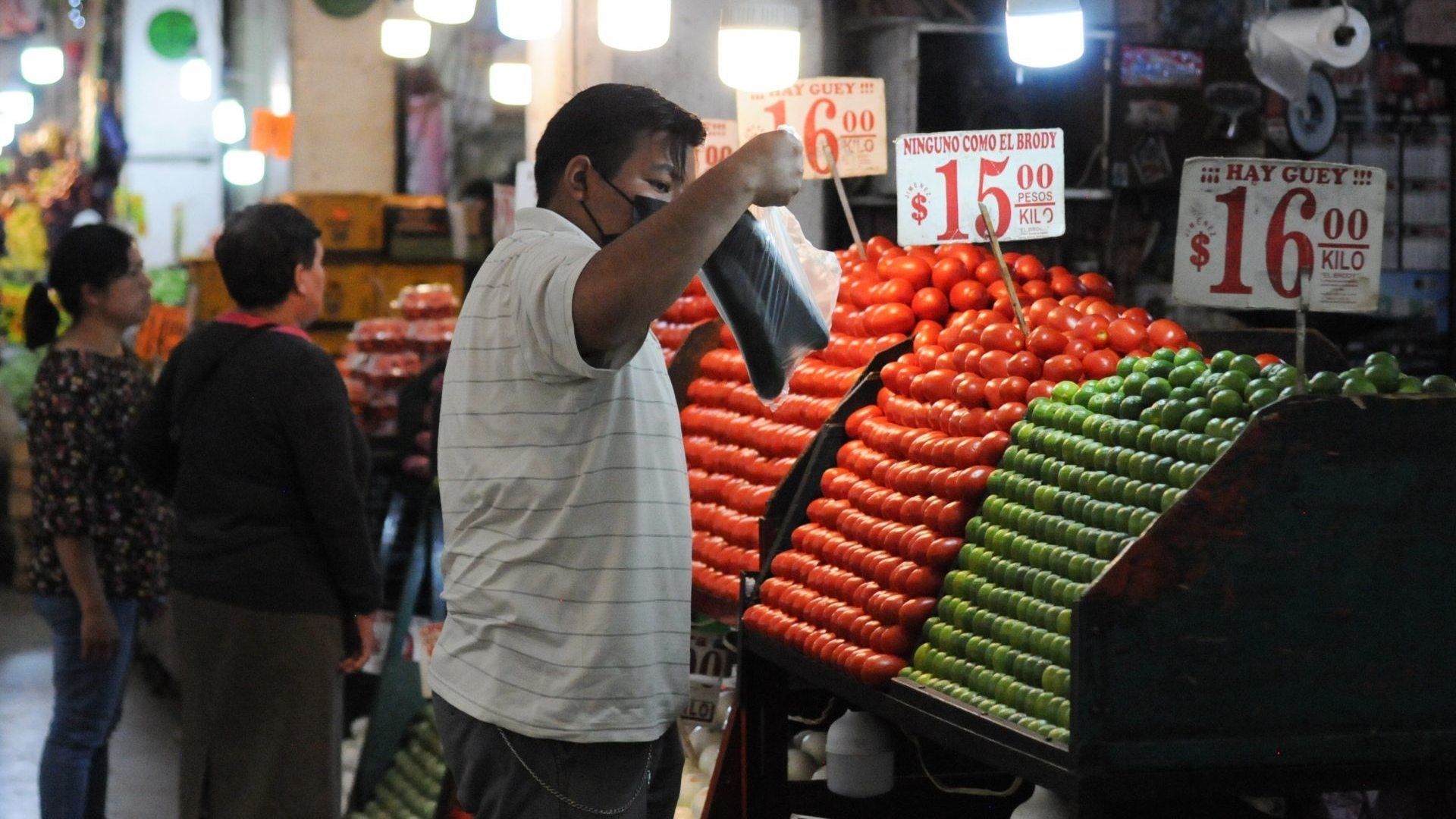 Una persona levanta una bolsa con pepinos frente a puesto de venta de verdura