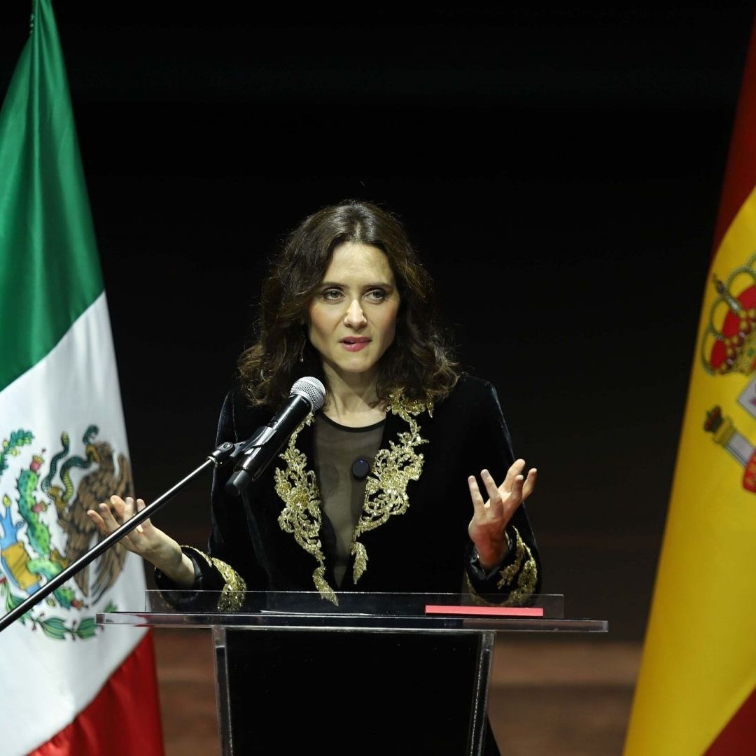 Isabel Díaz Ayuso habla frente a micrófonos con banderas de México y España de fondo