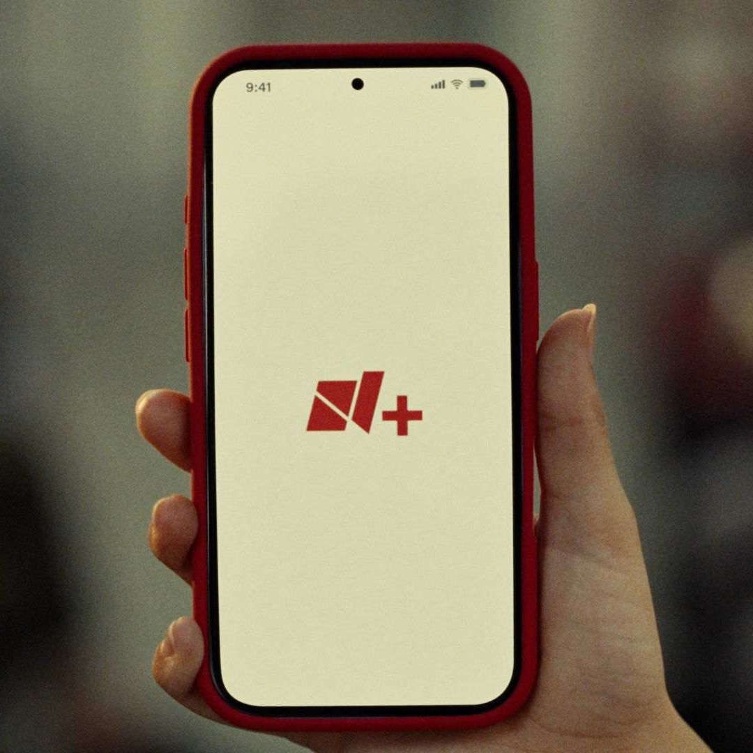 Mano sostiene celular con app y logo de Nmás. Nmás Lanza App en agosto 2026